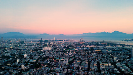 Aerial view capturing da nang cityscape, sea, and distant mountains bathed in soft pastel pink sunset light, revealing urban landscape against natural scenery
