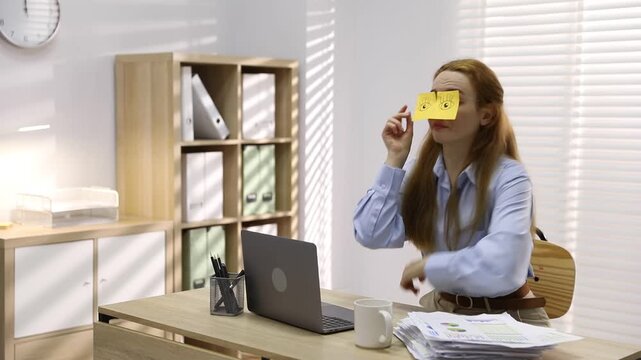 Sleepy woman yawning and putting on glasses with fake eyes drawn on sticky notes at workplace in office