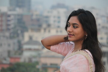 a portrait of a young woman in a light pink sari on a rooftop with a blurred city background!
