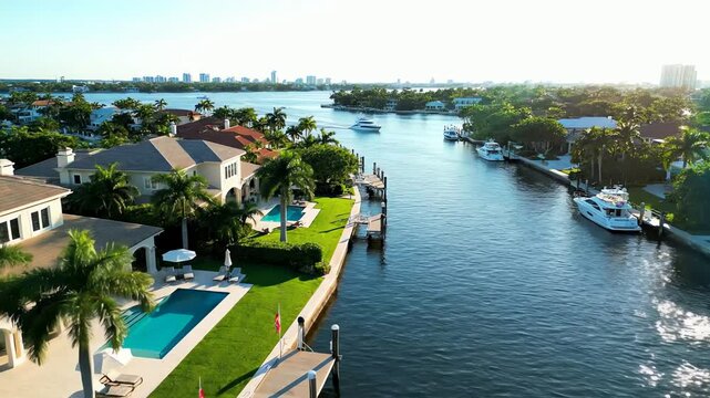 Stunning aerial view of luxurious waterfront mansions with private yachts along a serene canal with a city skyline in the distance.