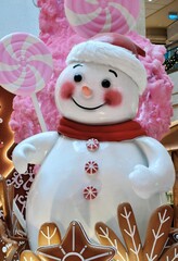 A close-up, vertical shot of a large, cute, white smiling snowman figure wearing a red scarf, positioned amongst oversized pink candy decorations and gingerbread cookies in a festive holiday display