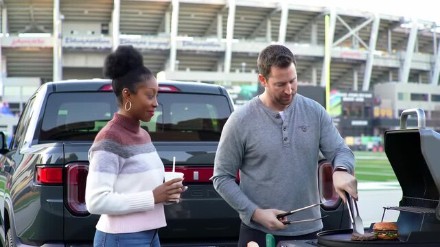 Happy diverse friends tailgating and grilling burgers on a pickup truck at a sports stadium, enjoying pre-game leisure.