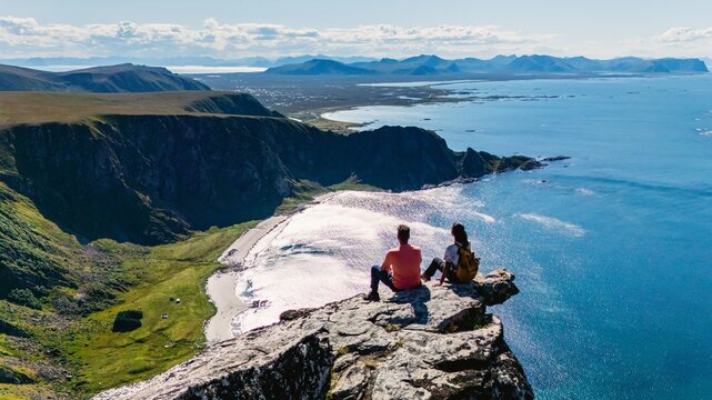 Two hikers enjoy a panoramic view from the edge of Matind Mountain, with Bleik Beach and the sea stretching into the horizon. The vibrant colors of nature create a stunning backdrop for adventure.