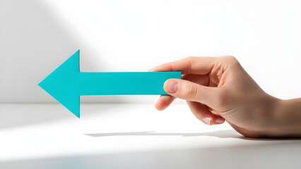 Woman's hand holding a turquoise arrow pointing left against a clean white background with strong directional light and shadow play
