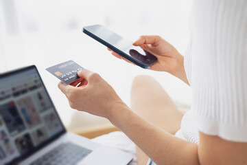 Woman hand holding a smartphone and credit card while shopping online with a laptop at home. Modern consumer lifestyle.