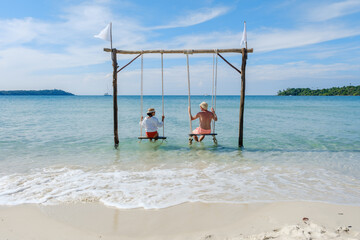 A diverse couple relaxing on swings by the serene beach of Koh Kood Island, Thailand. The calm waves and bright blue sky create a peaceful tropical atmosphere, perfect for unwinding.
