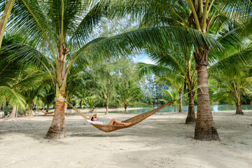 A peaceful scene of an Asian woman lounging in a hammock beneath tall palm trees on Koh Kood Island. Soft sandy beach and tranquil water create a tropical paradise in Thailand.
