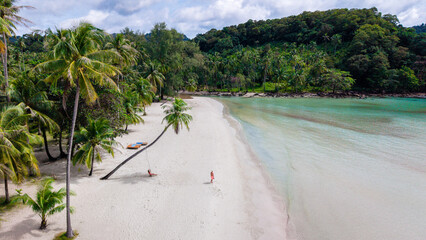 The serene beauty of Koh Kood Island in Thailand features soft sandy beaches, vibrant palm trees, and crystal-clear waters. A couple of men and a woman on a swing under a palm tree on the beach