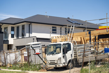 A residential construction site in a newly developed suburban estate in Australia, featuring a partially built house framed with timber, scaffolding, and safety fencing. A work truck is parked on site