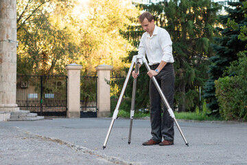 A man spreads and secures tripod legs outdoors, working carefully to prepare stable support for a...