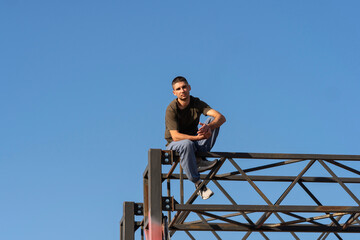 Close-up of a male freerunner in casual gear calmly balancing on a red-and-black metal frame under a cloudless sky, capturing strength, control, and urban athletic spirit