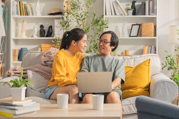 Asian married couple spends quality time together on a sofa in their living room at home, cheerfully using a laptop and smartphone, reflecting happiness and modern digital connection.