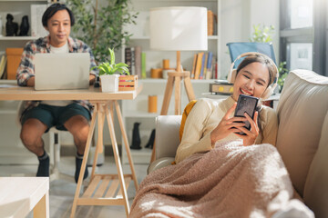 husband works remotely at home on his laptop while his wife lies cheerfully on the sofa, relaxing...
