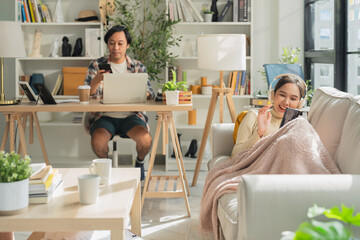 husband works remotely at home on his laptop while his wife lies cheerfully on the sofa, relaxing...