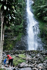 Obraz premium Father and son walking on rocky river near a tropical waterfall during outdoor adventure.