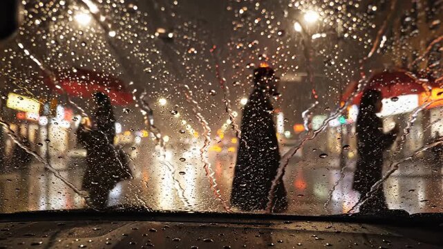 View from inside a car on a rainy street at night with raindrops on the windshield, blurred city lights reflecting off wet pavement.