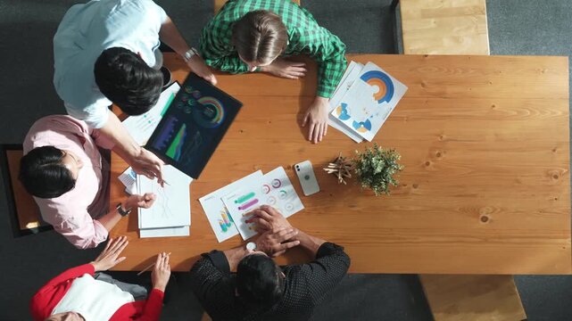 Top view of business man using tablet display increasing graph while diverse marketing people sharing idea or planing strategy at meeting table with stock market statistic scatter around. Convocation.