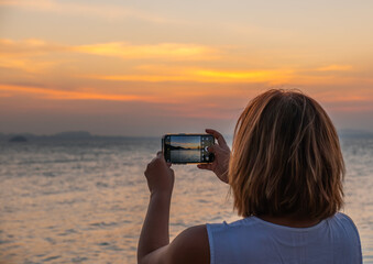 Asian woman capturing sunset scenery with smartphone—ideal for promoting scenic travel, digital nomad lifestyle, and nature photography tours.