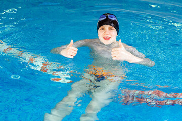 Preteen boy swimming in indoor pool during swim class and showing thumb up