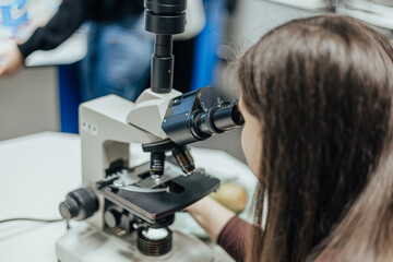 Young woman closely examining a sample through a laboratory microscope, adjusting the focus knobs with both hands in a classroom or research setting