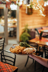 A server holds a plate with fries, chicken parmigiana, greens, and sauce in a warmly lit, decorated restaurant with plaid tablecloths and holiday lights