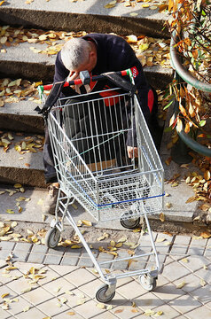 Vagabundo triste sin hogar en la calle con carro de la compra vac&iacute;o sentado en las escaleras en oto&ntilde;o