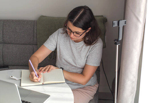 Adult woman writing notes in notebook while working at home, focused on planning and study, cozy home office lifestyle, remote work and productivity concept - Powered by Adobe