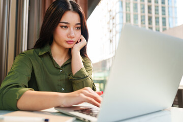 Young happy, beautiful, stylish, elegant Asian woman in green shirt working with computer laptop in luxury cafe. Digital technology, modern lifestyle, online learning and hybrid work