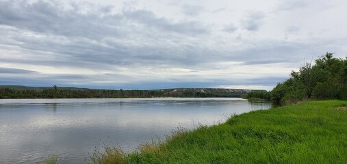 Fototapeta premium Panoramic river landscape with lush green grass meadow and forest bank under dramatic cloudy sky. Beautiful summer nature scenic view. Tranquil riverside environment background.