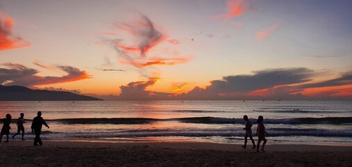 Fototapeta premium Dramatic sunrise over tropical beach with vibrant orange fire clouds and silhouettes of people walking along shore, beautiful morning sea landscape, summer vacation concept