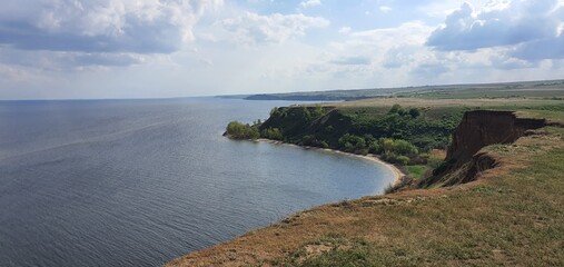 Scenic view of high clay cliffs and blue sea bay under cloudy sky. Coastal landscape with green hills and coastline. Beautiful nature panoramic view of shoreline and water.