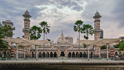 Sultan Abdul Samad Jamek Mosque, Kuala Lumpur, Malaysia. Moorish architecture with minarets and domes, viewed from the riverfront under a cloudy sky.