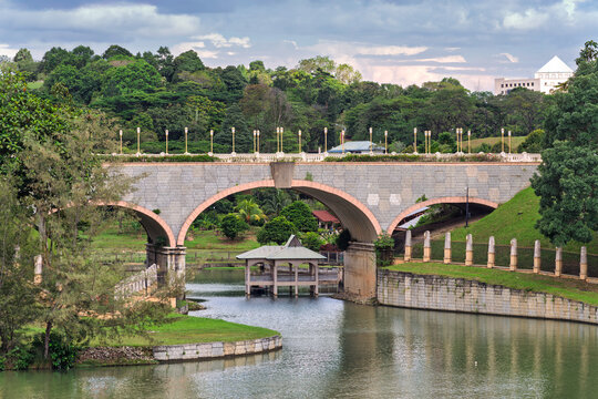 Seri Gemilang Bridge, Putrajaya, Malaysia. Arched stone bridge over water, surrounded by lush greenery, with a distant building under cloudy skies.