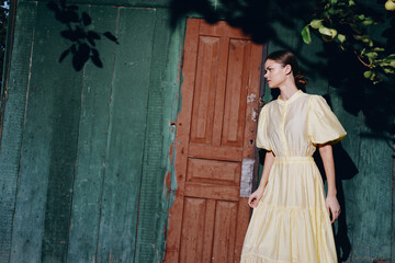 Woman in a pale yellow dress stands beside a weathered green wall and wooden door, an outdoor fashion portrait with soft light and thoughtful mood.