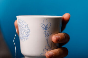 Close-up shot of hand holding a white teacup with a tea bag string against a blue background,...