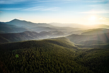 Aerial view of serene mountain landscape at sunset. Forested hills bathed in warm, golden light, creating tranquil atmosphere. Rolling hills stretch into the distance, covered in dense, lush greenery.