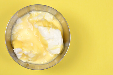 An overhead view shows a stainless steel bowl with snenokle dessert - meringues in vanilla custard - on a bright yellow background