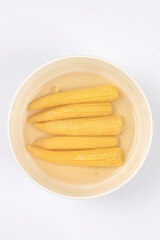 Five yellow canned baby corn cobs in a bowl of liquid on white background