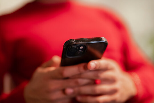 A person in a red shirt holds a black smartphone, focusing on the device's camera lens.