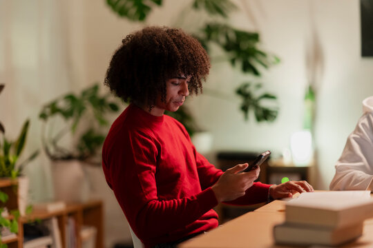 A young person with curly hair wearing a red sweater uses a smartphone while sitting at a table with books and plants in the background.