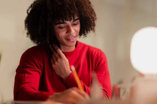 A smiling young man with curly hair is engaged in a phone conversation while simultaneously writing with a pencil. He wears a red sweater, working or studying.
