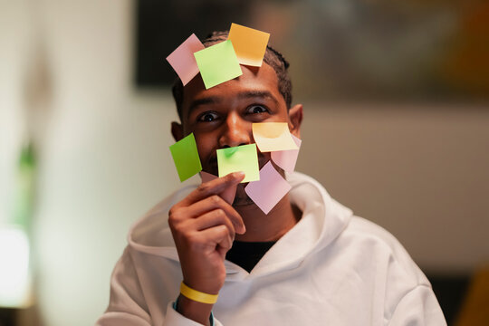 A man with colorful sticky notes on his face and head looks at the camera with a playful expression, holding one note near his mouth.