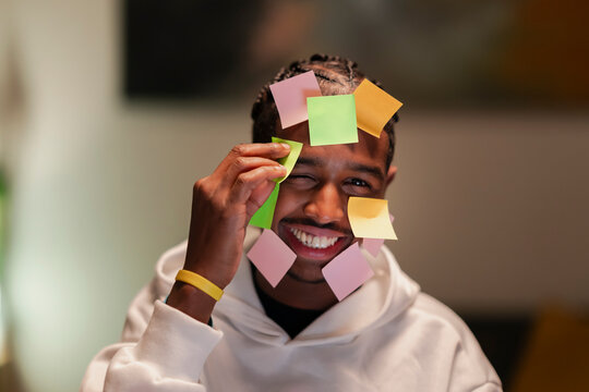 A young man with braided hair smiles and winks as he removes a colorful sticky note from his face, surrounded by many others.