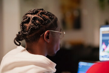 A young man with braided hair wears glasses and looks intently at a computer screen, engaged in work or study.