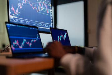 A person's hands are visible as they work on multiple computer screens displaying financial charts and data, suggesting analysis or trading.