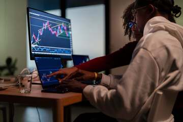 Two individuals collaborate, analyzing financial charts on multiple computer screens, indicating a focus on market trends and investment strategies.