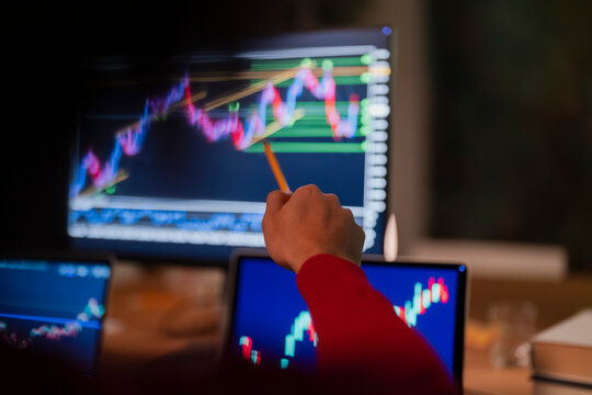A person in a red shirt uses a pencil to point at a stock market graph on a computer screen, with other screens showing similar data in the foreground.