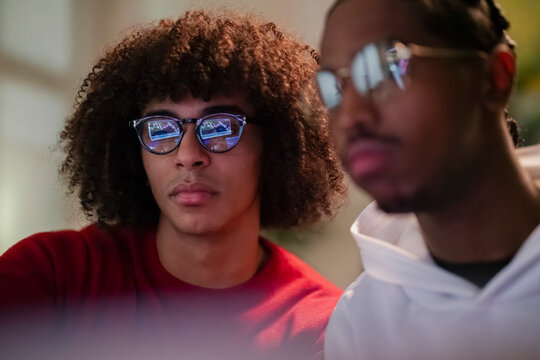 Two young men focus intently on a computer screen displaying financial charts, their reflections visible in the glasses of one.