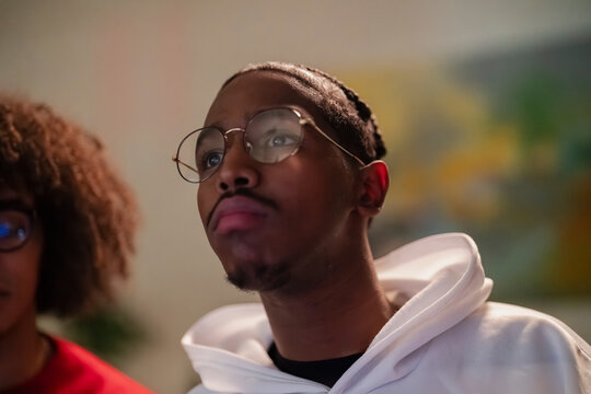 A young man wearing glasses and a white hoodie looks intently at something off-camera, with another person partially visible in the background.