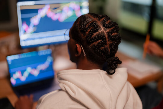 A person wearing glasses and a white hoodie analyzes financial charts on multiple computer screens, indicating a focus on trading or data analysis.
